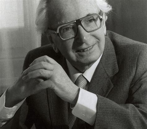 Black-and-white photo of an elderly man with white hair swept back, large rectangular glasses with thick dark frames, wearing a suit jacket, white shirt and striped tie, hands clasped, slight smile.